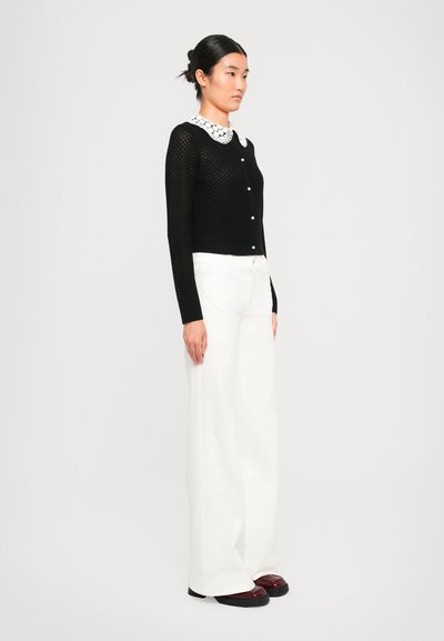 Black knitted cardigan with a textured pattern and white lace collar, paired with white wide-leg trousers and black shoes.