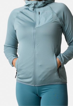 Femme portant une veste à capuche zippée bleu clair, les mains dans les poches latérales et des leggings bleus assortis.