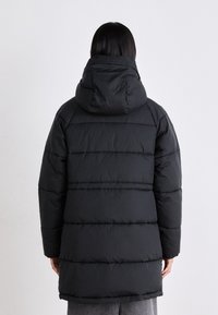 Osoba z prostymi, ciemnymi włosami, ubrana w długi, czarny, pikowany płaszcz zimowy z dużym kapturem, tyłem do kamery.