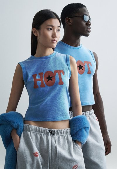 Top crop blu senza maniche con la scritta "HOT" e una grafica di una stella nera. Modelle che indossano pantaloni della tuta grigi, una con un accessorio peloso blu.