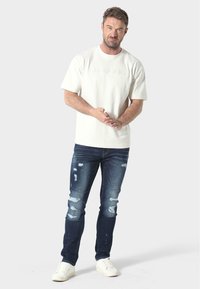 Camiseta oversized crema con letras en relieve, combinada con jeans oscuros desgastados y zapatillas blancas. Ropa casual simple y moderna.