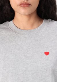 Gray crewneck sweatshirt with ribbed neckline, featuring a small red heart embroidered on the left chest. Soft, lightweight fabric.