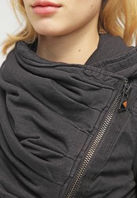 Gros plan d'une personne portant une veste gris foncé avec une fermeture éclair asymétrique et un grand col plié autour du cou.