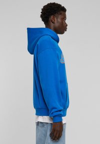 Sininen huppari, jossa on kengurupocket, pehmeä tekstuuri, ribbaus hihansuissa ja helmassa. Edessä monivärinen brodeerattu logo.