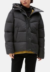 Czarna kurtka puchowa z kapturem, z zapięciem na zamek, pikowanym wzorem i bocznymi kieszeniami. Noszona na żółtym swetrze.