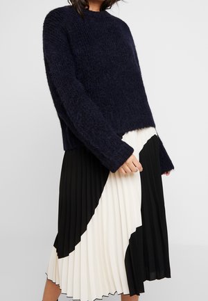 Kobieta w granatowym, puszystym swetrze, połączonym z czarno-białą, kolorową plisowaną spódnicą midi.