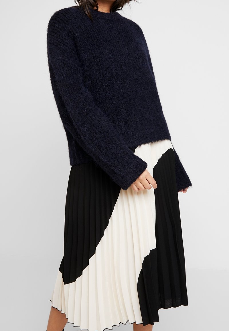 Kobieta w granatowym, puszystym swetrze, połączonym z czarno-białą, kolorową plisowaną spódnicą midi.