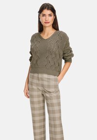 Olivgrüner gestrickter Pullover mit V-Ausschnitt und Spitzenmuster, kombiniert mit beigen karierten Weitbein-Hosen mit braunen Akzenten.