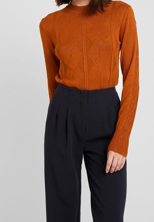 Femme portant un pull en maille texturée orange rouille à manches longues et un pantalon noir plissé taille haute sur fond gris.