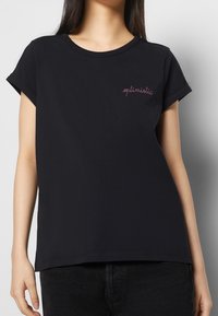 Svart t-shirt med kort ärm, tillverkad av mjuk bomull, med en simpel söm som säger "optimistisk" i rosa. Rak nederkant och avslappnad passform.