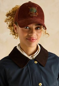 Căciulă din catifea bordou cu un logo brodat circular, purtată cu o haină albastră închisă cu guler din catifea maronie și butoane din alamă.