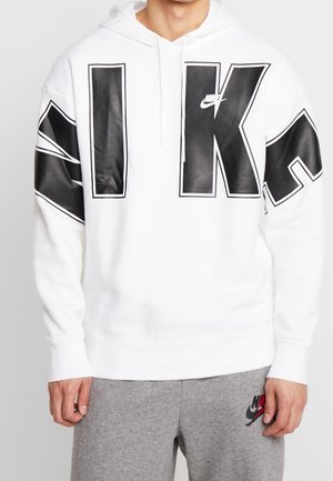 Osoba nosząca białą bluzę z kapturem Nike z dużymi czarnymi literami na piersi oraz szare spodnie dresowe z małym logo Nike na udzie.