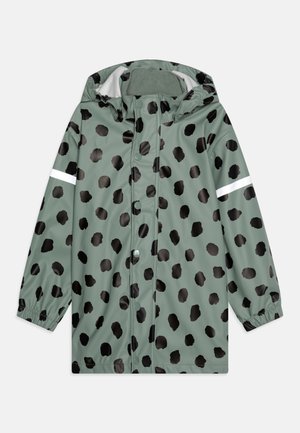 Veste imperméable verte avec des pois noirs irréguliers. Équipée d'une capuche, de boutons-pression, de poignets élastiques et d'une bande réfléchissante sur la manche.