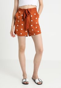 Pantaloni corti arancioni con pois bianchi, dotati di vita legata e tasche laterali. Realizzati in un tessuto leggero con una superficie liscia.