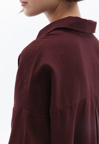 Camicia di cotone borgogna con colletto, vestibilità rilassata e dettagli arricciati lungo la cucitura posteriore. Tessuto morbido e liscio.