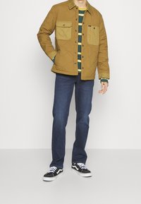 Braune gesteppte Jacke mit grünen Akzenttaschen über einem grün-gelb gestreiften Hemd, kombiniert mit dunkelblauen Jeans und schwarzen Turnschuhen.