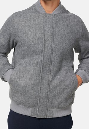 Mann trägt eine graue Woll-Bomberjacke mit gerippten Bündchen und Saum, Hände in den Taschen, vor einem schlichten hellen Hintergrund.