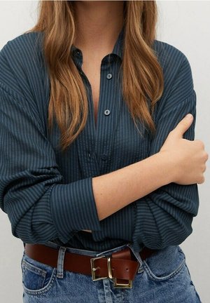 Femme aux longs cheveux châtain clair portant une chemise à boutons rayée bleu foncé rentrée dans un jean bleu avec une ceinture marron, les bras croisés.