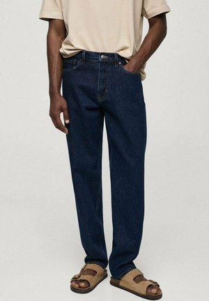 Voksen iført mørkeblå jeans med lige ben, beige kortærmet skjorte og brune åbne sandaler, stående med den ene hånd i lommen.
