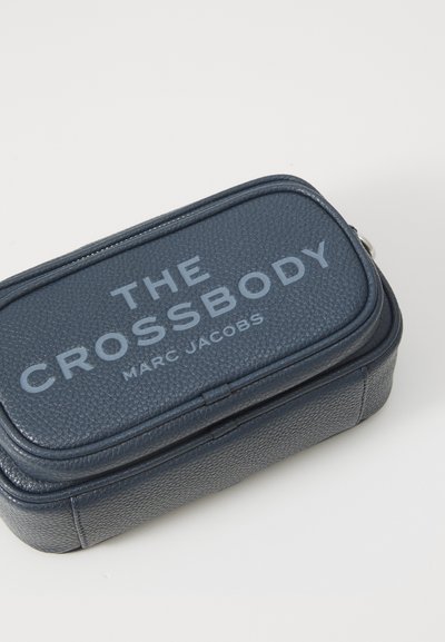 Geantă din piele albastru marin, tip crossbody, cu o suprafață texturată, având textul embosat "THE CROSSBODY" și "MARC JACOBS" în gri. Închidere cu fermoar.