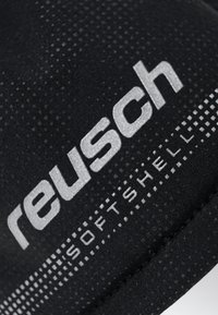 Schwarzer Reusch Softshell-Stoff mit strukturierter Oberfläche und reflektierender silberner Markenkennzeichnung. Zeigt gepunktete Muster entlang des Randes.