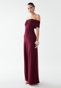 Bordeauxroter schulterfreier Jumpsuit mit Raffung, weit geschnittenen Beinen und glattem Stoff. Kombiniert mit einem massiven goldenen Armband und Riemchensandalen.