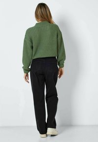 Olivgrüner Strickpullover mit strukturiertem Muster, kombiniert mit hoch taillierter schwarzer Jeans. Das Model zeigt seinen Rücken zur Kamera und trägt cremefarbene Schuhe.