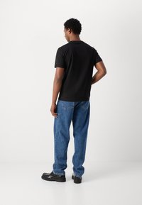 Camiseta negra de manga corta, de corte relajado, combinada con jeans de mezclilla azules, con corte recto y costuras visibles en el bolsillo trasero. Zapatos negros.