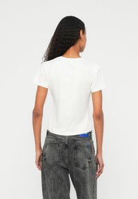 Γυναίκα με σγουρά μαλλιά που φοράει απλό λευκό κοντομάνικο μπλουζάκι και ψηλόμεσα ξεθωριασμένα μαύρα τζιν, εμφανιζόμενη από πίσω.