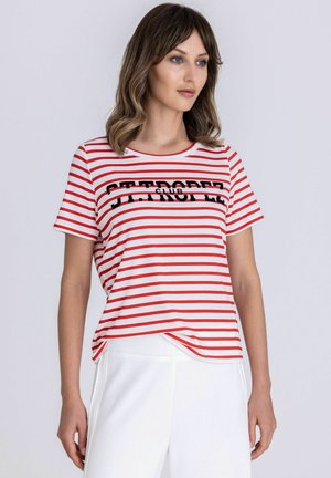 Vrouw draagt een rood-wit gestreept shirt met korte mouwen met de tekst "ST. TROPEZ CLUB", gecombineerd met een witte broek, staand tegen een effen achtergrond.