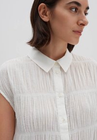 Una camicia bianca con colletto e maniche corte, realizzata in un tessuto testurizzato e plissettato, con chiusura a bottoni sul davanti. Dettagli di cuciture orizzontali sottili.
