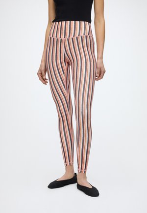 Γυναίκα που φοράει ψηλόμεσα κολάν με κάθετες ρίγες σε ροζ, μαύρο και πορτοκαλί, συνδυασμένα με μαύρο αμάνικο τοπ και μαύρα μπαλαρίνες.