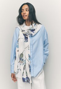 Chemise bleu clair à boutons associée à une écharpe blanche ornée de motifs floraux bleus et verts, mettant en valeur un tissu doux et un style drapé.