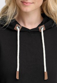 Sweatshirt à capuche noir en tissu texturé, avec des cordons de tirage crème et des accents en cuir marron au niveau du col. Vue en gros plan.