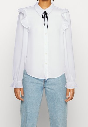Biała bluzka z długim rękawem z falbankami na ramionach i mankietach, zapinana z przodu na guziki, z czarną wstążką przy kołnierzu, noszona z jasnoniebieskimi dżinsami.