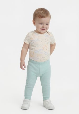 Μικρό παιδί που στέκεται, φοράει κρέμ πουκάμισο με ζωάκια, ανοιχτό μπλε ριμπ παντελόνι και λευκά αθλητικά, χαμογελάει και κοιτάζει προς τα κάτω.