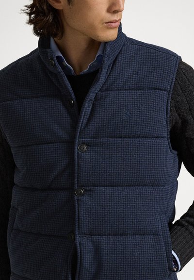 Μπλε σκούφος αμάνικο μπουφάν με υφή με σχέδιο, διαθέτει όρθιο γιακά, κουμπιά στο μπροστινό μέρος και πλαϊνές τσέπες, φορεμένο πάνω από μαύρο πουλόβερ.