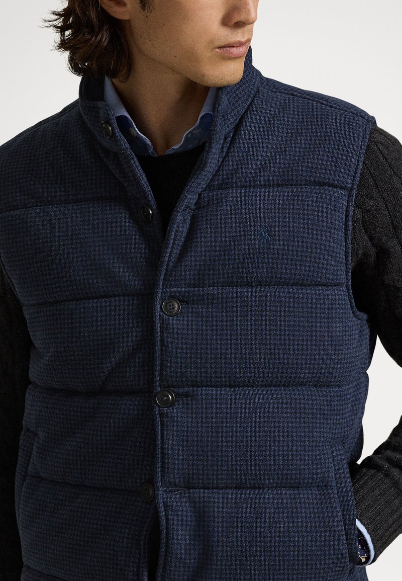 Marineblaue Steppweste mit gemustertem Texturdesign, mit Stehkragen, vorderen Knöpfen und Seitentaschen, über einem schwarzen Pullover getragen.