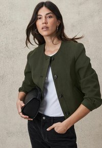 Olive green jacket with a round neckline, front buttons, and three-quarter sleeves, paired with a white top and black clutch bag.