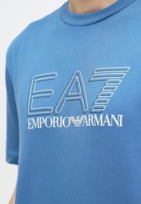 Sinine särk, millel on valgete ja ääristatud tähtedega "EA7 Emporio Armani" logo rinnal, kantud inimese poolt.