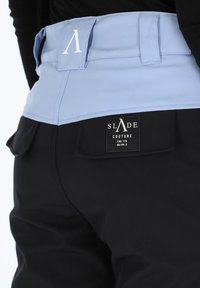 Black pants with a light blue waistband, featuring a logo patch labeled "SLADE COUTURE." The fabric appears smooth with a functional pocket.