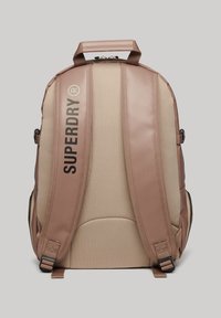 Zaino in rosa chiaro e beige, realizzato in tessuto testurizzato. Presenta spallacci imbottiti, dettagli con logo e una maniglia superiore.