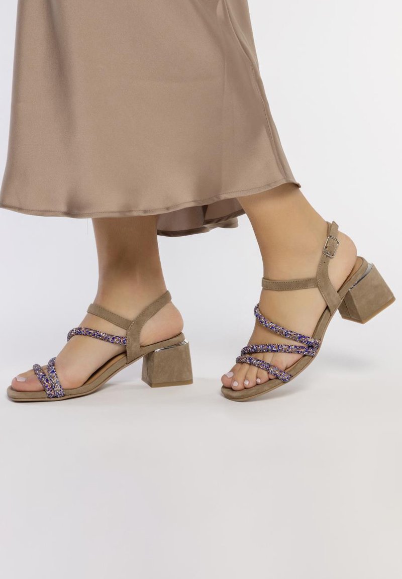 Sandalias de tacón de ante beige con tiras metálicas moradas entrelazadas, tacón cuadrado y diseño de espalda abierta, exhibidas sobre un fondo neutro.