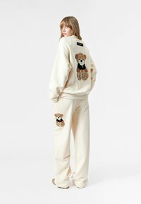 Conjunto de sudadera y pantalones de color crema que presenta apliques de osito de peluche en la parte trasera y lateral, con detalles texturizados y un corte relajado.