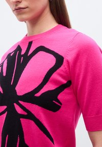 Γυναίκα που φοράει ένα φωτεινό ροζ πουλόβερ με κοντά μανίκια, με μεγάλο αφαιρετικό μαύρο σχέδιο στο στήθος και τον ώμο.