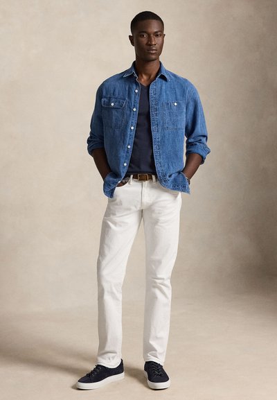 Fiatal férfi áll, kezét zsebében tartva, kék farmeringet, fekete pólót, fehér nadrágot, barna övet és fekete tornacipőt viselve.