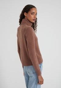 Giovane donna che indossa un maglione a collo alto marrone e jeans azzurri chiari, che guarda sopra la spalla contro uno sfondo semplice.