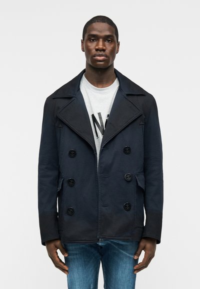 Homme portant un manteau croisé bleu marine sur un t-shirt gris clair avec un graphique et un jean bleu, debout devant un fond blanc uni.