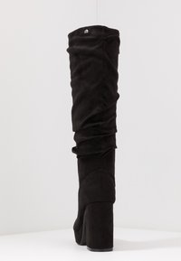Bota negra por encima de la rodilla, hecha de suave ante, con un diseño arrugado, tacón grueso alto y un detalle de logo en la parte superior.