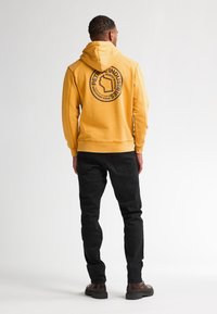 Felpa con cappuccio gialla con un logo grafico circolare nero sul retro, abbinata a pantaloni neri e stivali marroni.